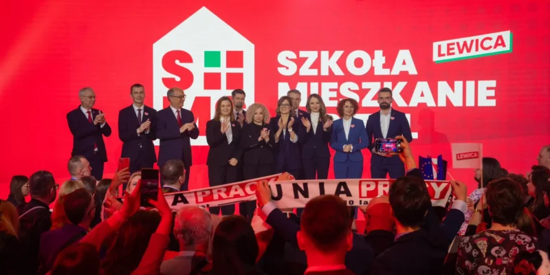 Grupa polityków Lewicy stoi na scenie podczas konwencji samorządowej, przed czerwonym tłem z napisem „SZKOŁA MIESZKANIE” i logo Lewicy, wśród wiwatującego tłumu i osób robiących zdjęcia.