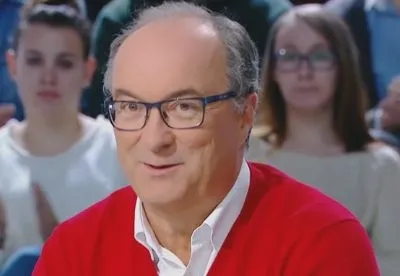 Mężczyzna w czerwonym swetrze i okularach uśmiecha się podczas rozmowy w telewizyjnym studiu, w tle widać słuchaczy.