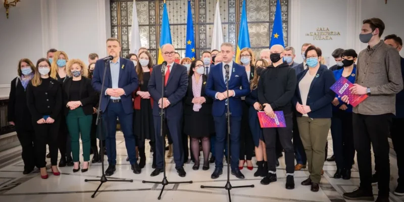 Grupa osób w maseczkach stoi przed mikrofonami, podczas konferencji prasowej w eleganckim pomieszczeniu z flagami Polski i Ukrainy w tle.