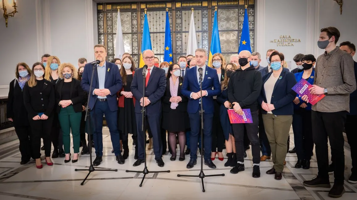 Grupa osób w maseczkach stoi przed mikrofonami, podczas konferencji prasowej w eleganckim pomieszczeniu z flagami Polski i Ukrainy w tle.