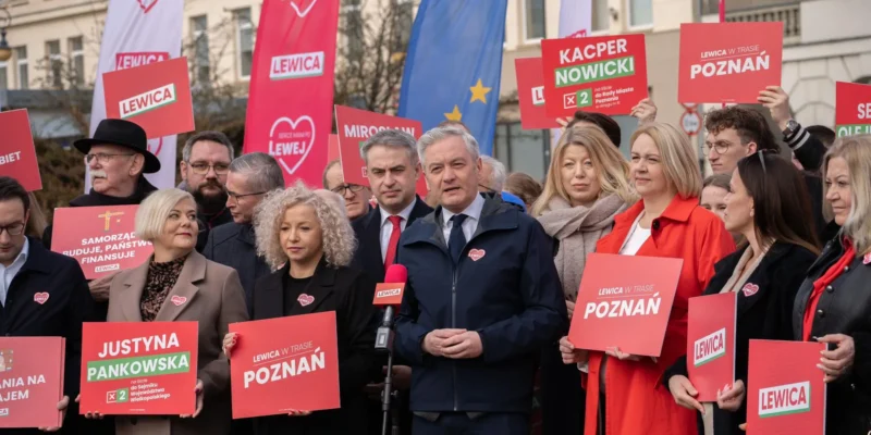Grupa zwolenników Lewicy na plenerowym wydarzeniu trzyma czerwone plakaty z nazwami miast i słucha mówcy przy mikrofonie.