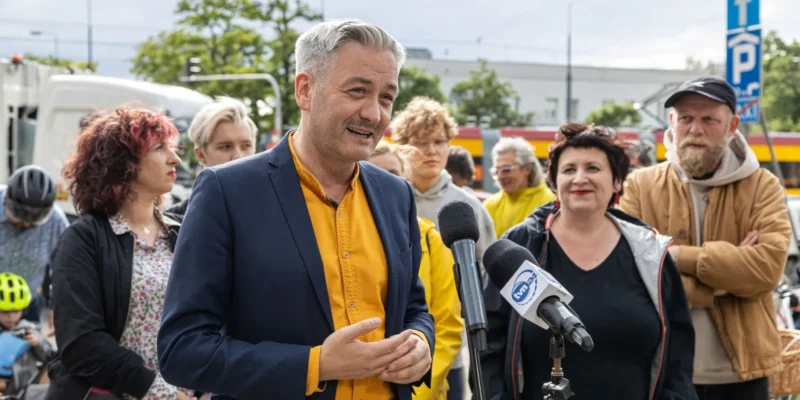 Na zdjęciu widać mężczyznę w garniturze, który przemawia do zgromadzonej grupy ludzi, a w tle dostrzegalne są rowery i miejskie otoczenie.