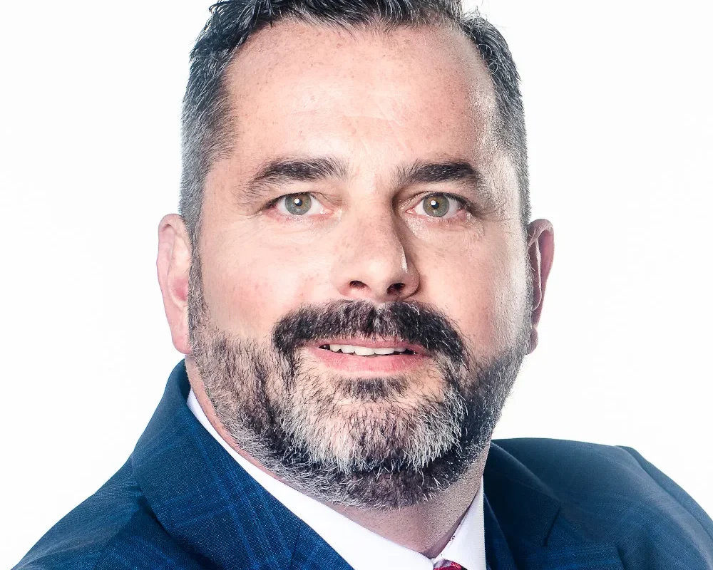 Mężczyzna w granatowym garniturze z czerwonym krawatem, z brodą i uśmiechem, patrzy w obiektyw na białym tle.