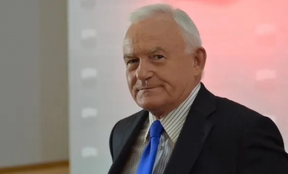 Starszy mężczyzna w ciemnym garniturze i niebieskim krawacie patrzy poważnie w aparat na tle rozmytego jasnoczerwonego ekranu.