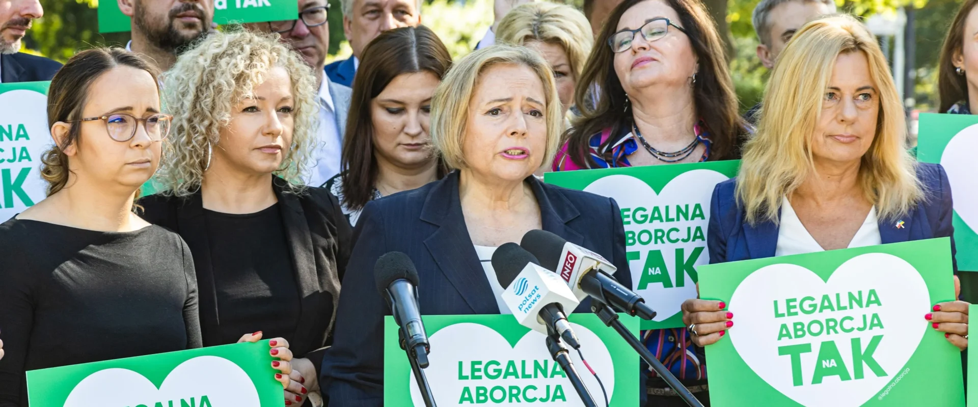 Grupa osób na demonstracji z zielonymi plakatami „Legalna aborcja TAK”, wśród nich kobieta w garniturze przemawia do mikrofonów.