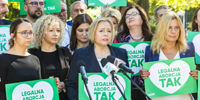 Grupa osób na demonstracji z zielonymi plakatami „Legalna aborcja TAK”, wśród nich kobieta w garniturze przemawia do mikrofonów.