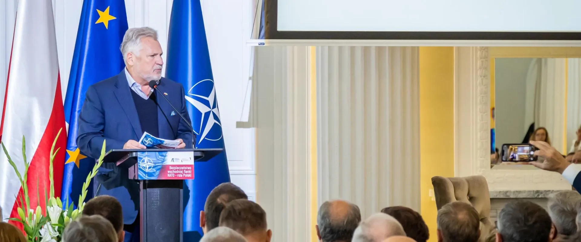 Na zdjęciu widać mężczyznę przemawiającego na konferencji nt. bezpieczeństwa w związku z NATO, z flagami Polski oraz Unii Europejskiej w tle.