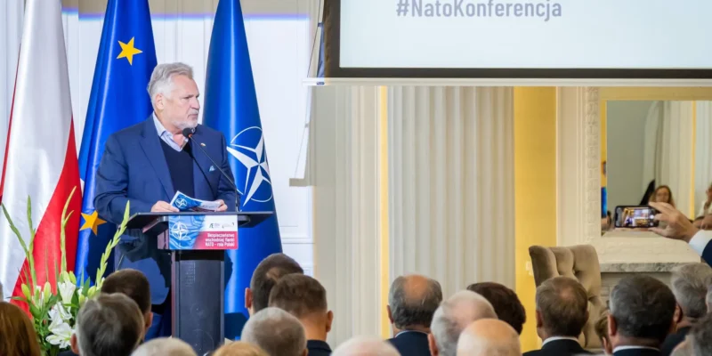 Na zdjęciu widać mężczyznę przemawiającego na konferencji nt. bezpieczeństwa w związku z NATO, z flagami Polski oraz Unii Europejskiej w tle.