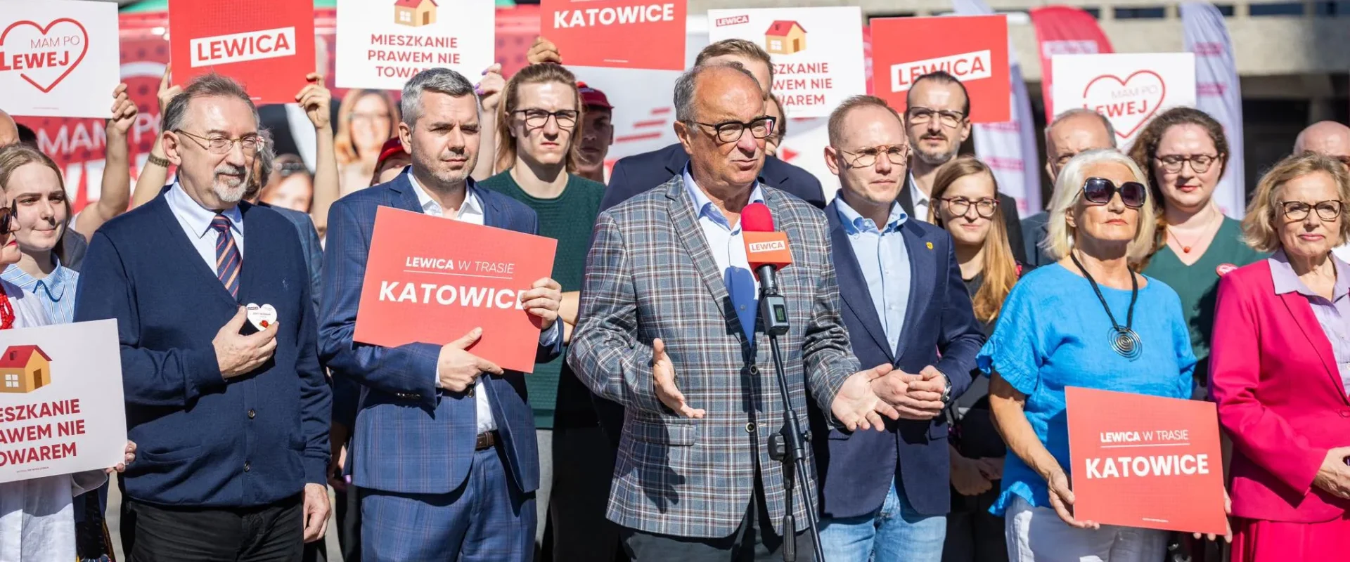 Grupa polityków Lewicy na wydarzeniu w Katowicach, trzymają plakaty z napisem Lewica i Katowice, podczas gdy jeden mężczyzna przemawia do mikrofonu.