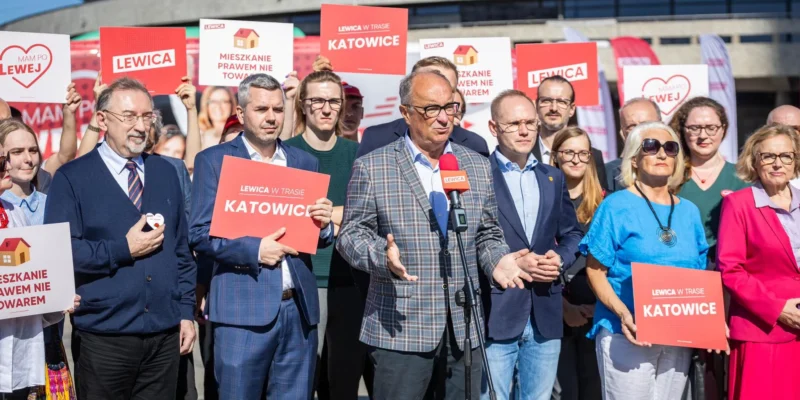 Grupa polityków Lewicy na wydarzeniu w Katowicach, trzymają plakaty z napisem Lewica i Katowice, podczas gdy jeden mężczyzna przemawia do mikrofonu.