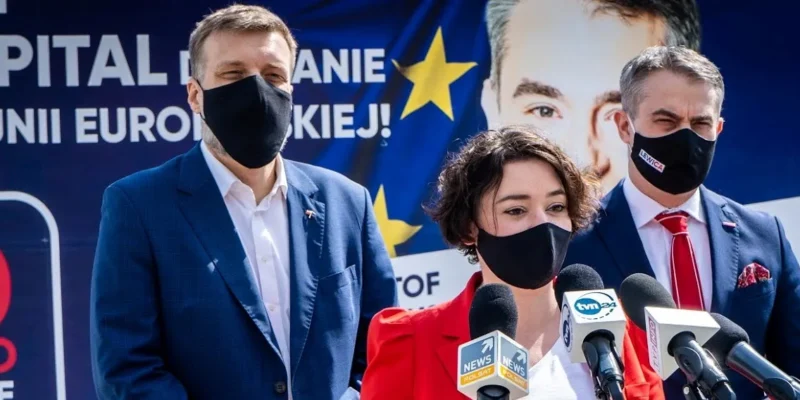 Trzy osoby w maseczkach stoją przed banerem Lewicy z mikrofonami podczas konferencji prasowej, a kobieta w czerwonej marynarce jest na pierwszym planie.