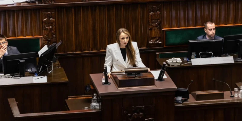 Kobieta w białym garniturze przemawia z mównicy w sali plenarnej parlamentu.