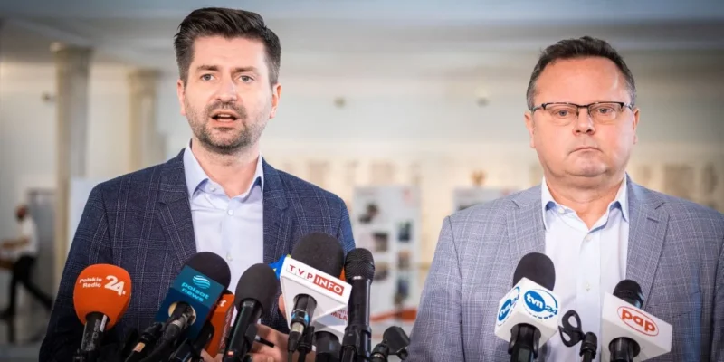 Dwóch mężczyzn w garniturach przemawia podczas konferencji prasowej, stojąc przed zestawem mikrofonów w sprawie uchodźców w Polsce.