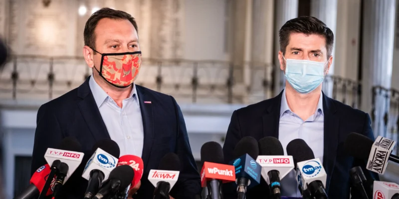 Dwóch mężczyzn w garniturach, w maseczkach ochronnych, prowadzi konferencję prasową dotyczącą Krajowego Planu Odbudowy, stojąc obok mikrofonów.