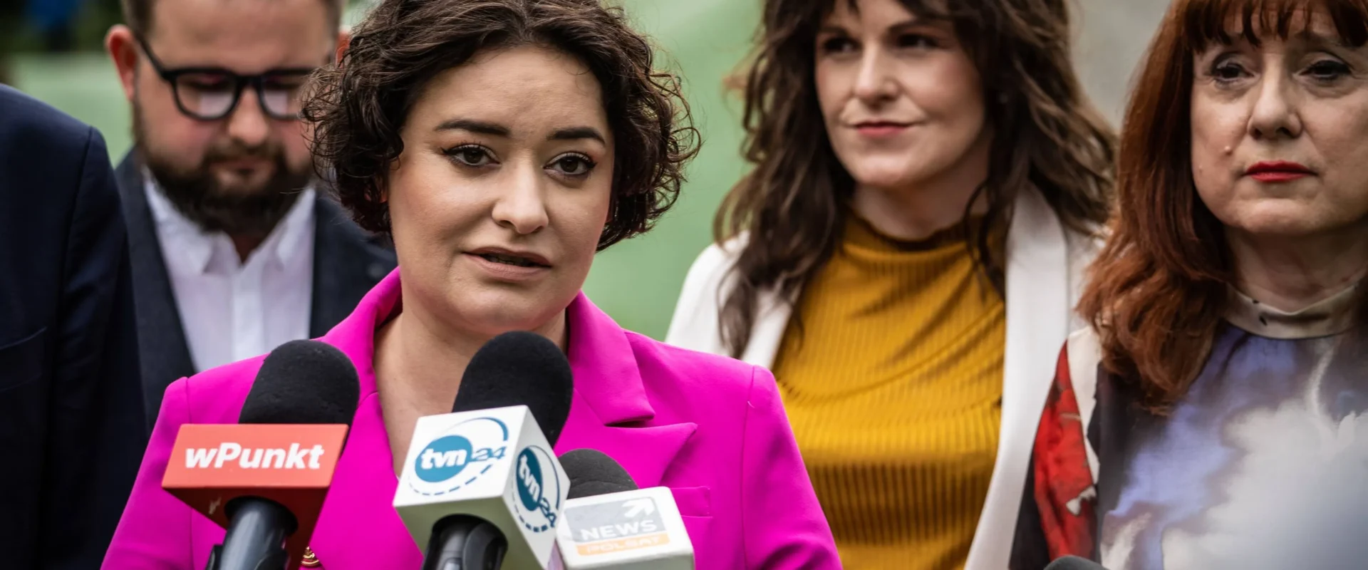 Kobieta w jaskraworóżowym garniturze przemawia do mediów przed zestawem mikrofonów podczas konferencji prasowej, otoczona innymi osobami.