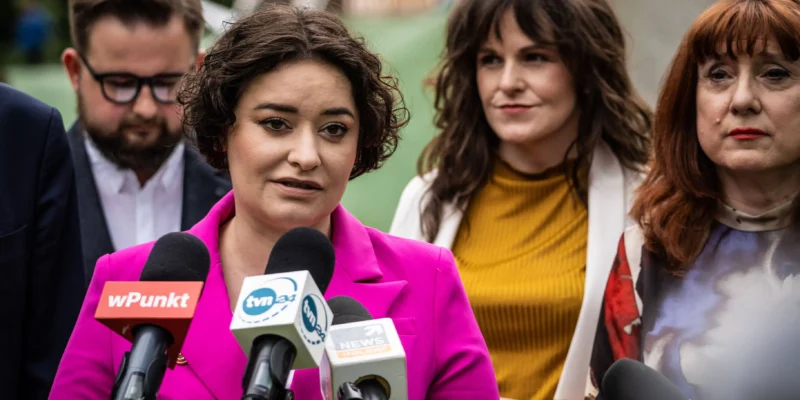 Kobieta w jaskraworóżowym garniturze przemawia do mediów przed zestawem mikrofonów podczas konferencji prasowej, otoczona innymi osobami.