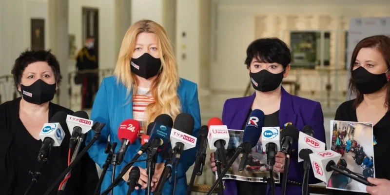 Cztery kobiety w maskach ochronnych stoją przed mikrofonami podczas konferencji prasowej w budynku.