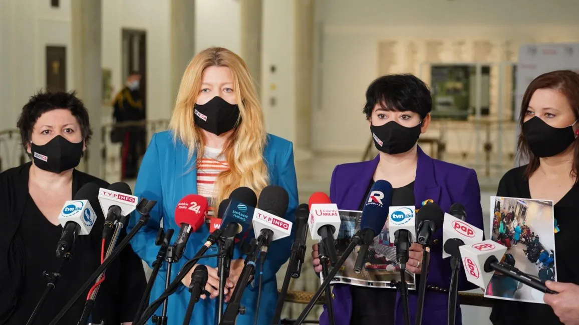 Cztery kobiety w maskach ochronnych stoją przed mikrofonami podczas konferencji prasowej w budynku.