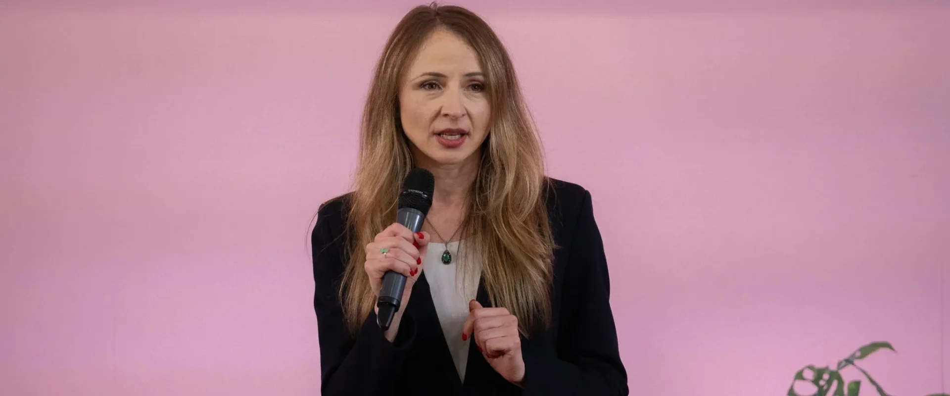 Kobieta z długimi blond włosami, ubrana w czarny żakiet, trzyma mikrofon i przemawia na różowym tle.