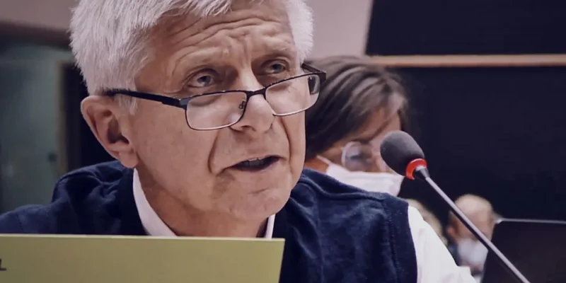 Starszy mężczyzna z siwymi włosami i okularami siedzi przy mikrofonie podczas konferencji, trzyma żółtą kartkę.