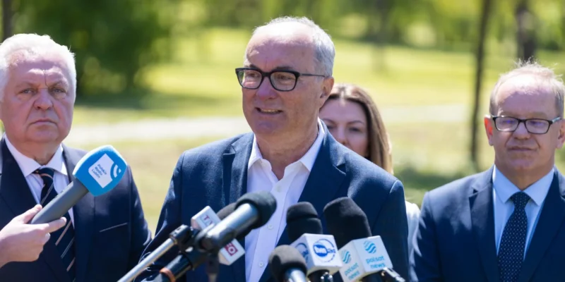 Na zdjęciu widoczny jest mężczyzna w okularach, który mówi do mikrofonów, a w tle stoją inni mężczyźni oraz kobieta, otoczeni zielonymi drzewami.