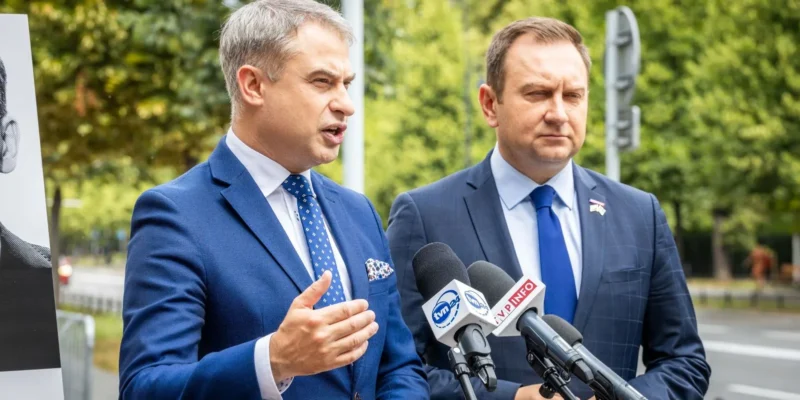 Dwóch mężczyzn w garniturach przemawia na konferencji prasowej na zewnątrz, stojąc obok mikrofonów ustawionych na statywach.