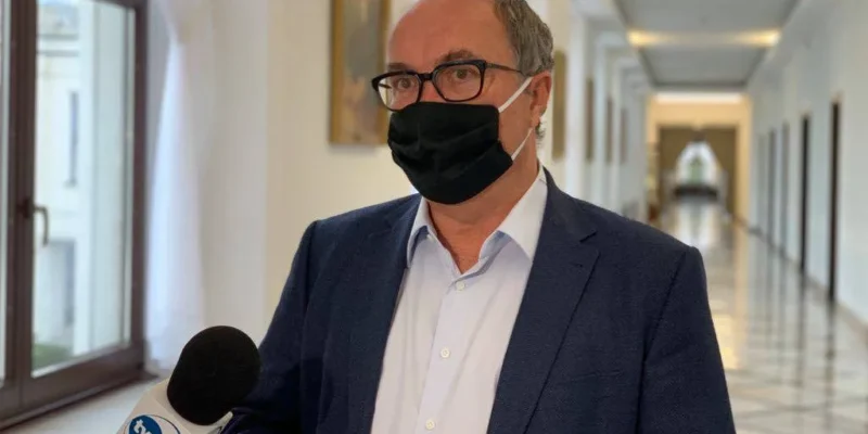 Osoba w granatowym garniturze, białej koszuli i maseczce ochronnej stoi w jasnym, długim korytarzu z oknami z lewej strony, trzymając mikrofon z logo TVN.