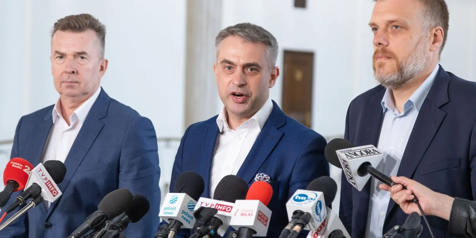 Na zdjęciu trzech mężczyzn w garniturach stoi przed mikrofonami podczas konferencji prasowej.