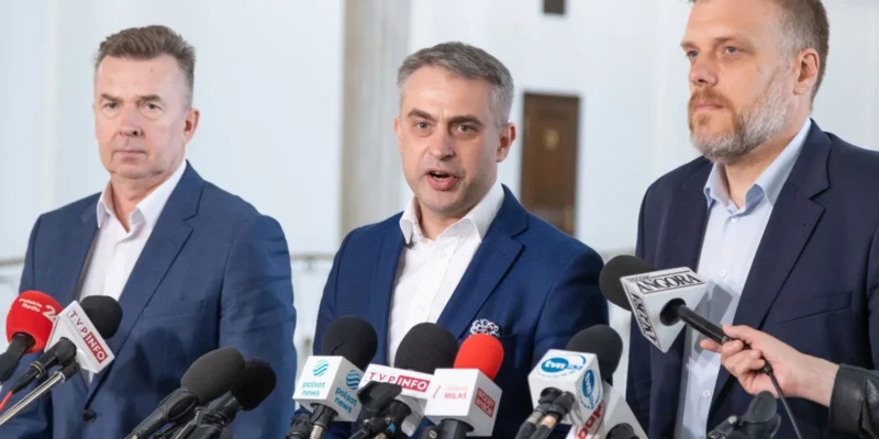 Na zdjęciu trzech mężczyzn w garniturach stoi przed mikrofonami podczas konferencji prasowej.