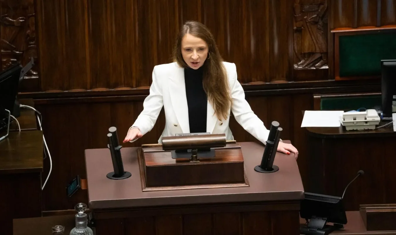 Kobieta w białej marynarce przemawia z mównicy w sali plenarnej parlamentu.
