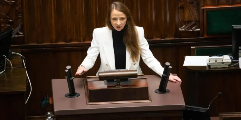 Kobieta w białej marynarce przemawia z mównicy w sali plenarnej parlamentu.