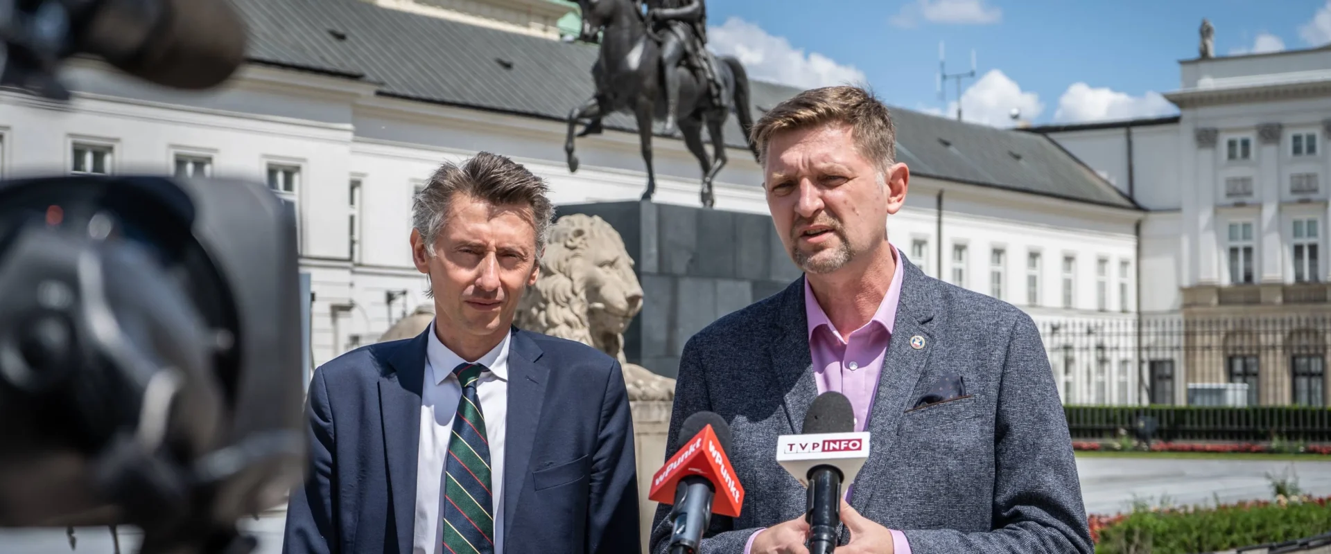 Dwóch mężczyzn w garniturach udziela wywiadu przed kamerami na placu z posągiem konia w tle i zabytkowymi budynkami.