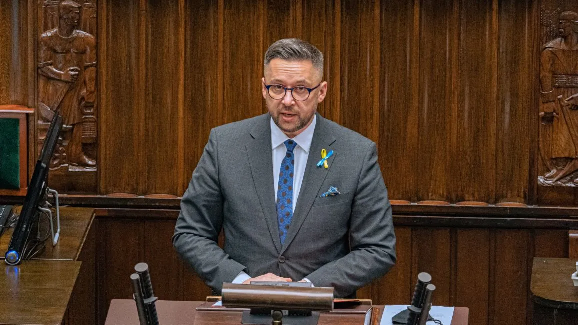 Mężczyzna w garniturze i okularach przemawia z mównicy podczas debaty nad ustawą o obronie ojczyzny; na klapie ma żółto-niebieską wstążkę, a w tle widoczne są drewniane dekoracje sali.