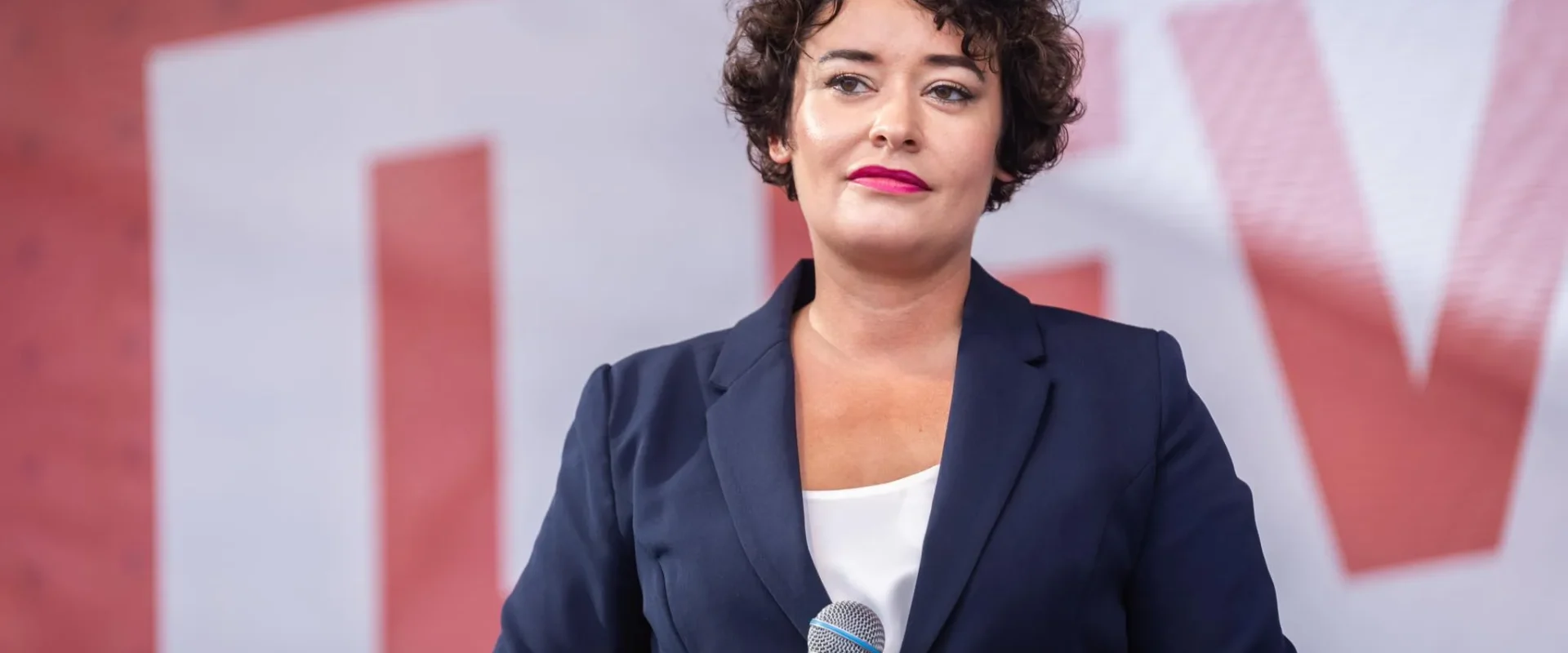Kobieta o krótkich kręconych włosach, ubrana w granatowy żakiet i białą bluzkę, trzyma mikrofon podczas przemówienia.