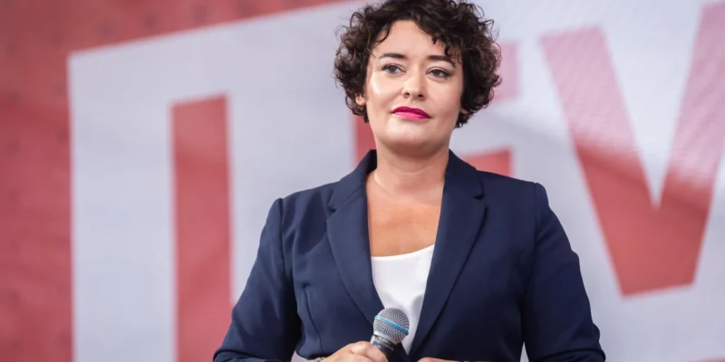 Kobieta o krótkich kręconych włosach, ubrana w granatowy żakiet i białą bluzkę, trzyma mikrofon podczas przemówienia.