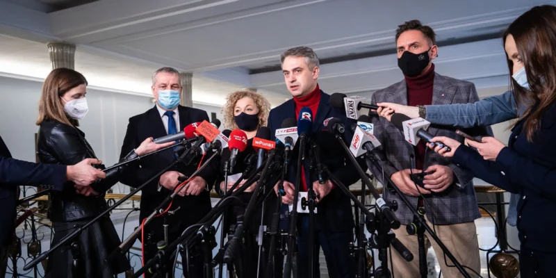 Grupa polityków i dziennikarzy podczas konferencji prasowej, otoczona licznymi mikrofonami i kamerami, wszyscy w maseczkach.