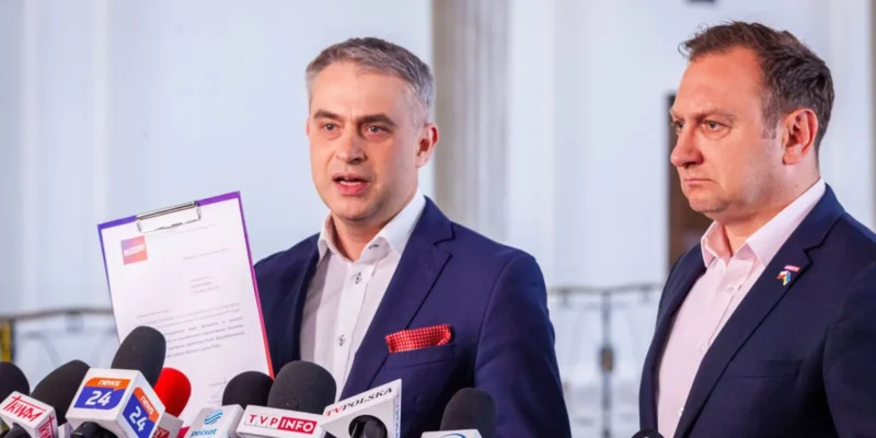 Dwóch mężczyzn w garniturach podczas konferencji prasowej na temat afery w NCBiR; jeden z nich trzyma teczkę z dokumentem, otoczony przez liczne mikrofony mediów.