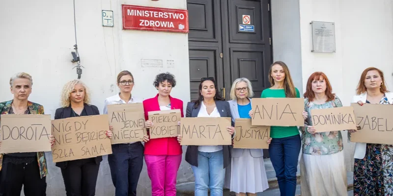 Grupa kobiet stoi przed wejściem do budynku Ministerstwa Zdrowia, trzymając kartonowe tabliczki z imionami i protestuje przeciwko drakońskiemu prawu antyaborcyjnemu w Polsce.