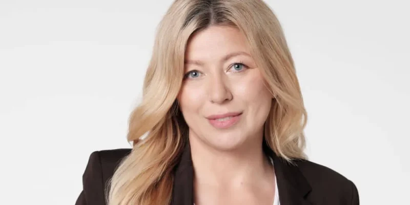 Portret uśmiechniętej blondynki w czarnym żakiecie i białej bluzce na jasnym tle.