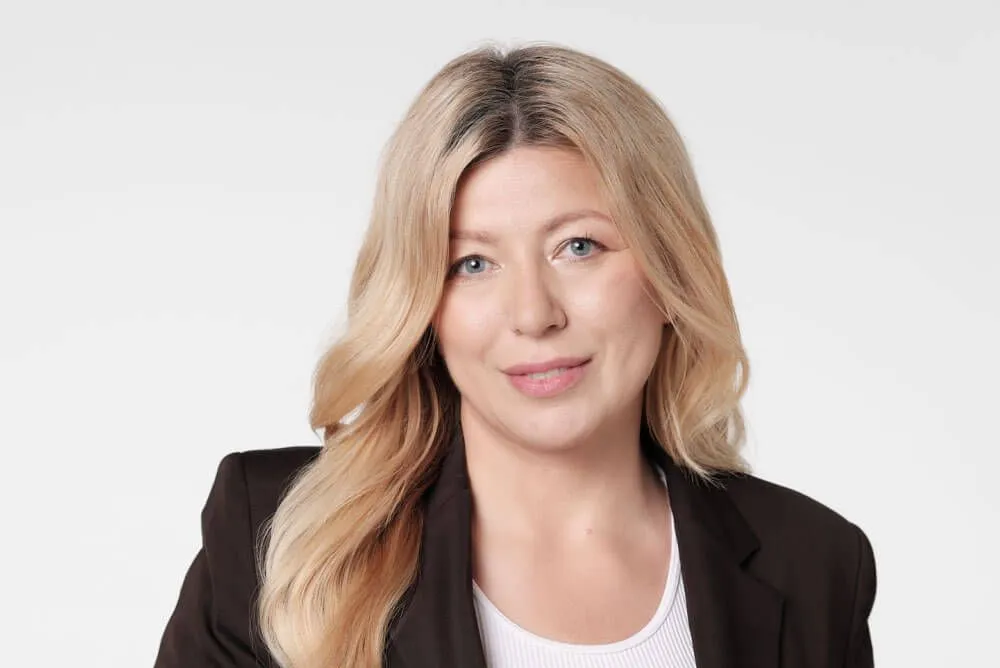 Portret uśmiechniętej blondynki w czarnym żakiecie i białej bluzce na jasnym tle.