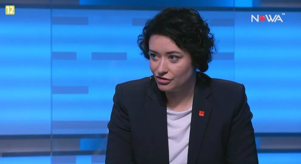 Kobieta w ciemnej marynarce i białej koszulce mówiąca w studiu telewizyjnym z niebieskim tłem i logiem stacji w prawym górnym rogu.