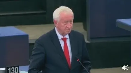 Mężczyzna w granatowym garniturze i czerwonym krawacie, o siwych włosach, przemawia przy mównicy na sali obrad.