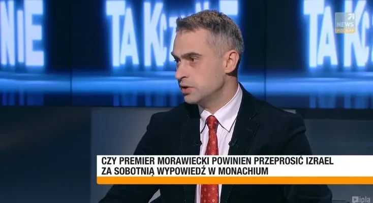 Mężczyzna w garniturze i czerwonym krawacie przemawia w studiu telewizyjnym, a na pasku pojawia się pytanie, czy premier Morawiecki powinien przeprosić Izrael za wypowiedź w Monachium.