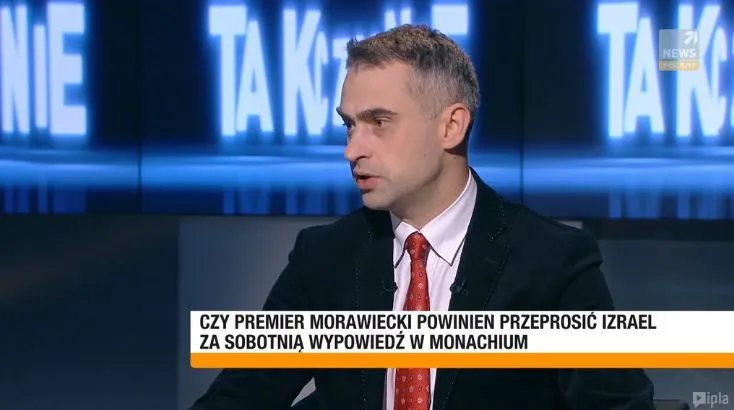 Mężczyzna w garniturze i czerwonym krawacie przemawia w studiu telewizyjnym, a na pasku pojawia się pytanie, czy premier Morawiecki powinien przeprosić Izrael za wypowiedź w Monachium.
