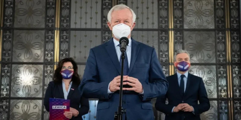 Polityk w granatowym garniturze i białej masce przemawia do mikrofonu podczas konferencji; w tle dwie osoby w garniturach i maseczkach, jedna z nich trzyma folder z napisem LEWICA.