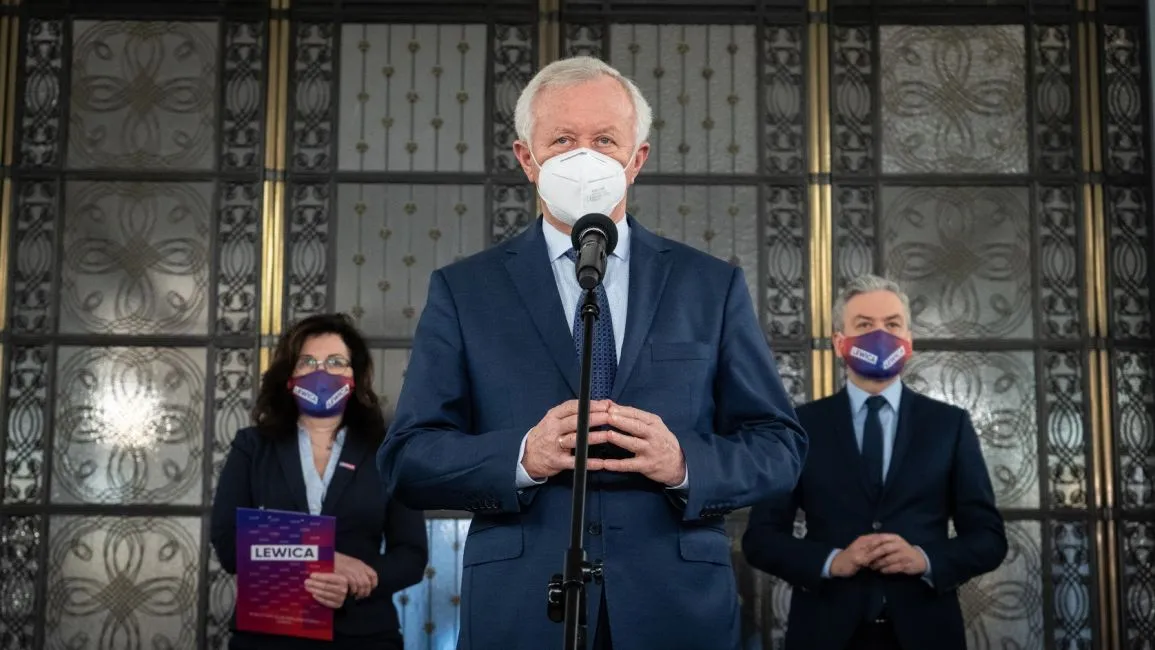 Polityk w granatowym garniturze i białej masce przemawia do mikrofonu podczas konferencji; w tle dwie osoby w garniturach i maseczkach, jedna z nich trzyma folder z napisem LEWICA.