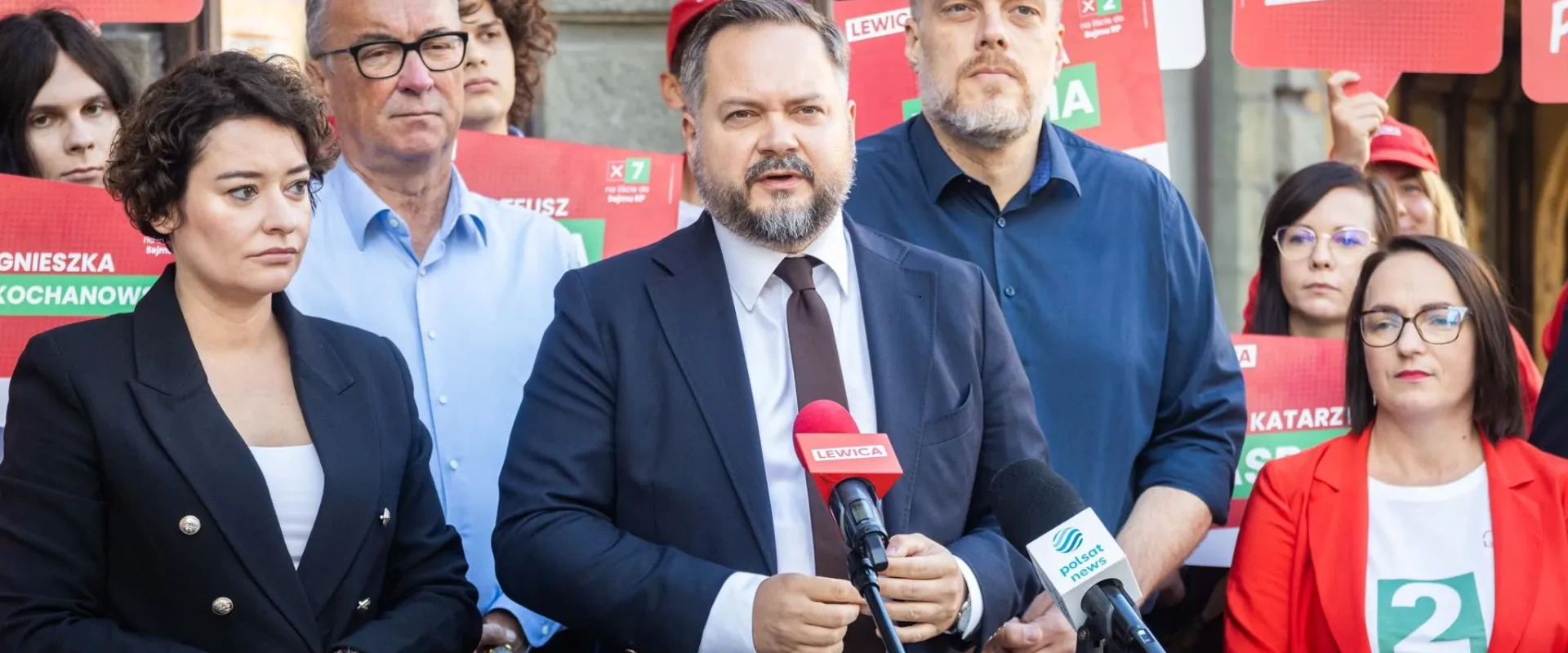 Mężczyzna w garniturze przemawia na konferencji prasowej Lewicy, otoczony przez zwolenników trzymających czerwone plakaty i mikrofony z logami partii.