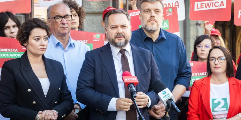 Mężczyzna w garniturze przemawia na konferencji prasowej Lewicy, otoczony przez zwolenników trzymających czerwone plakaty i mikrofony z logami partii.