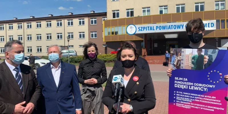 Grupa osób w maseczkach stoi przed wejściem do szpitala powiatowego, a jedna kobieta przemawia do mikrofonu obok plakatu promującego Fundusz Odbudowy.
