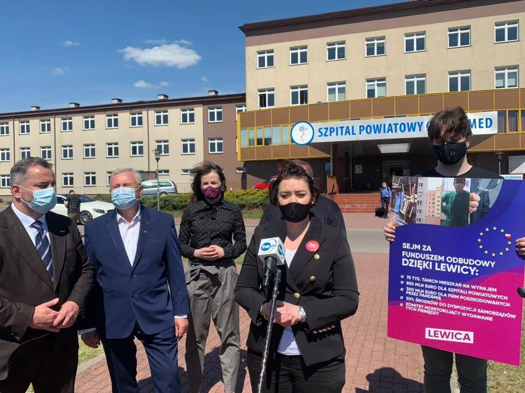 Grupa osób w maseczkach stoi przed wejściem do szpitala powiatowego, a jedna kobieta przemawia do mikrofonu obok plakatu promującego Fundusz Odbudowy.